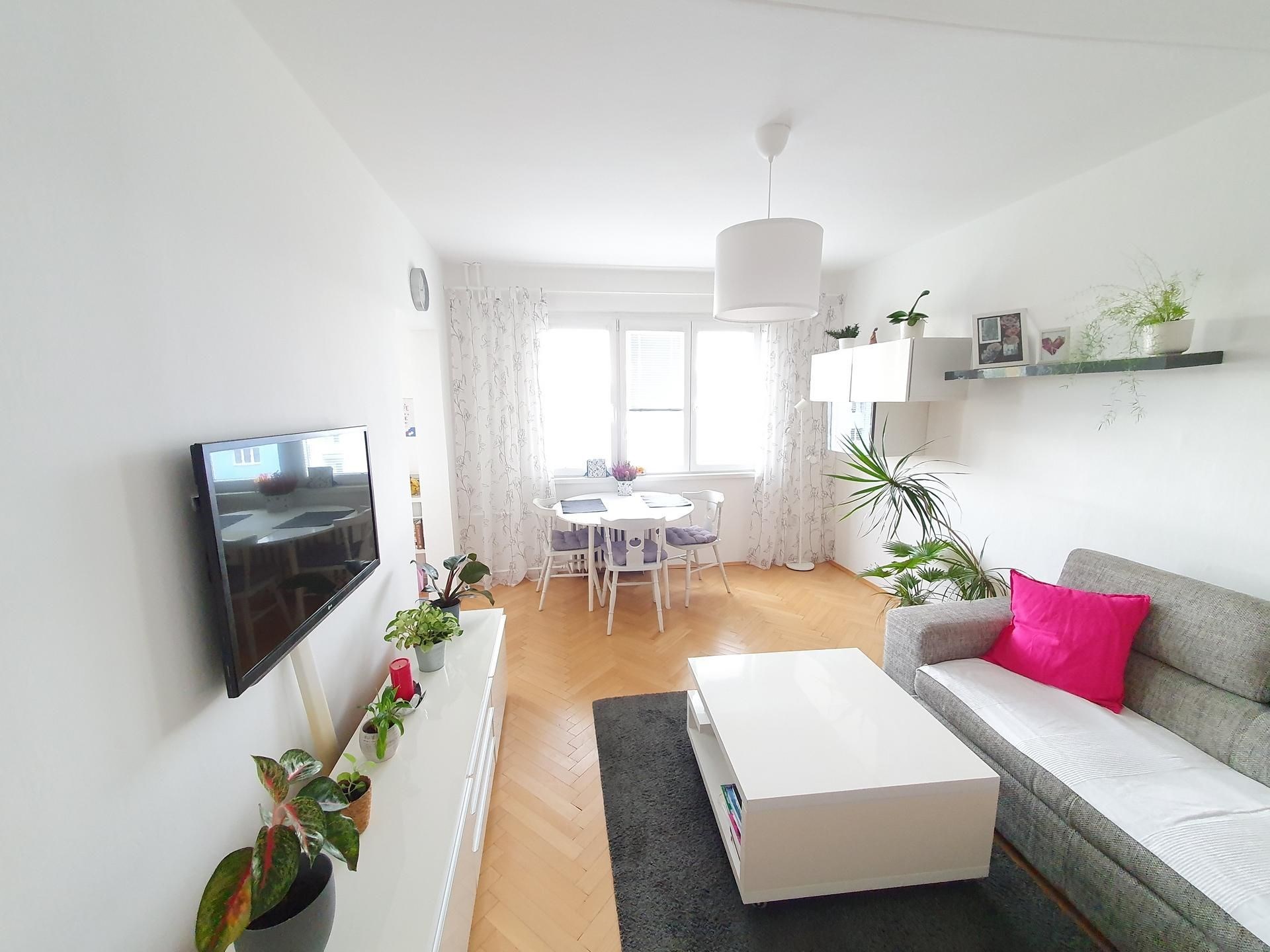 Světle zařízený obývací pokoj s parketovou podlahou, velkými okny a moderním nábytkem.