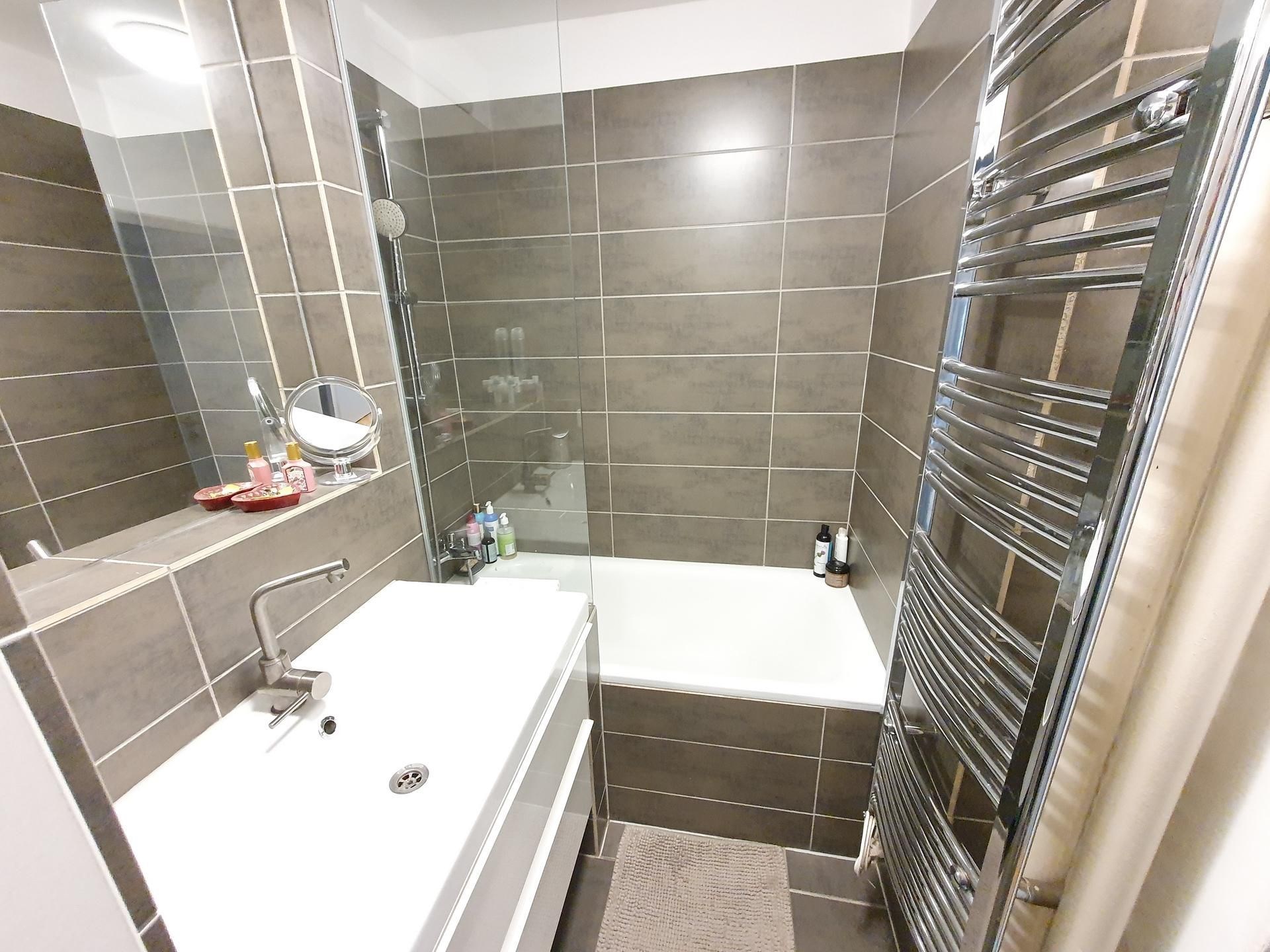 Moderní koupelna s dlažbou, skleněnou sprchovou zástěnou a chromovaným radiátorem, nabízí elegantní a praktický design.