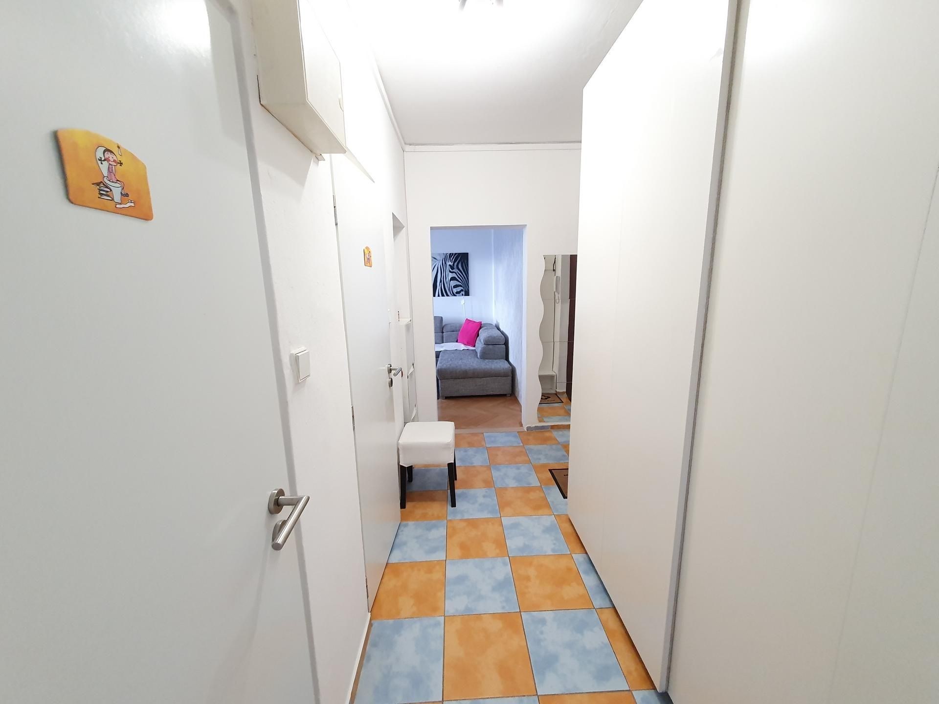 Úzká chodba s dlažbou a vestavěnými skříněmi, moderní světlý interiér nabízí výhled do obývacího pokoje s pohovkou.