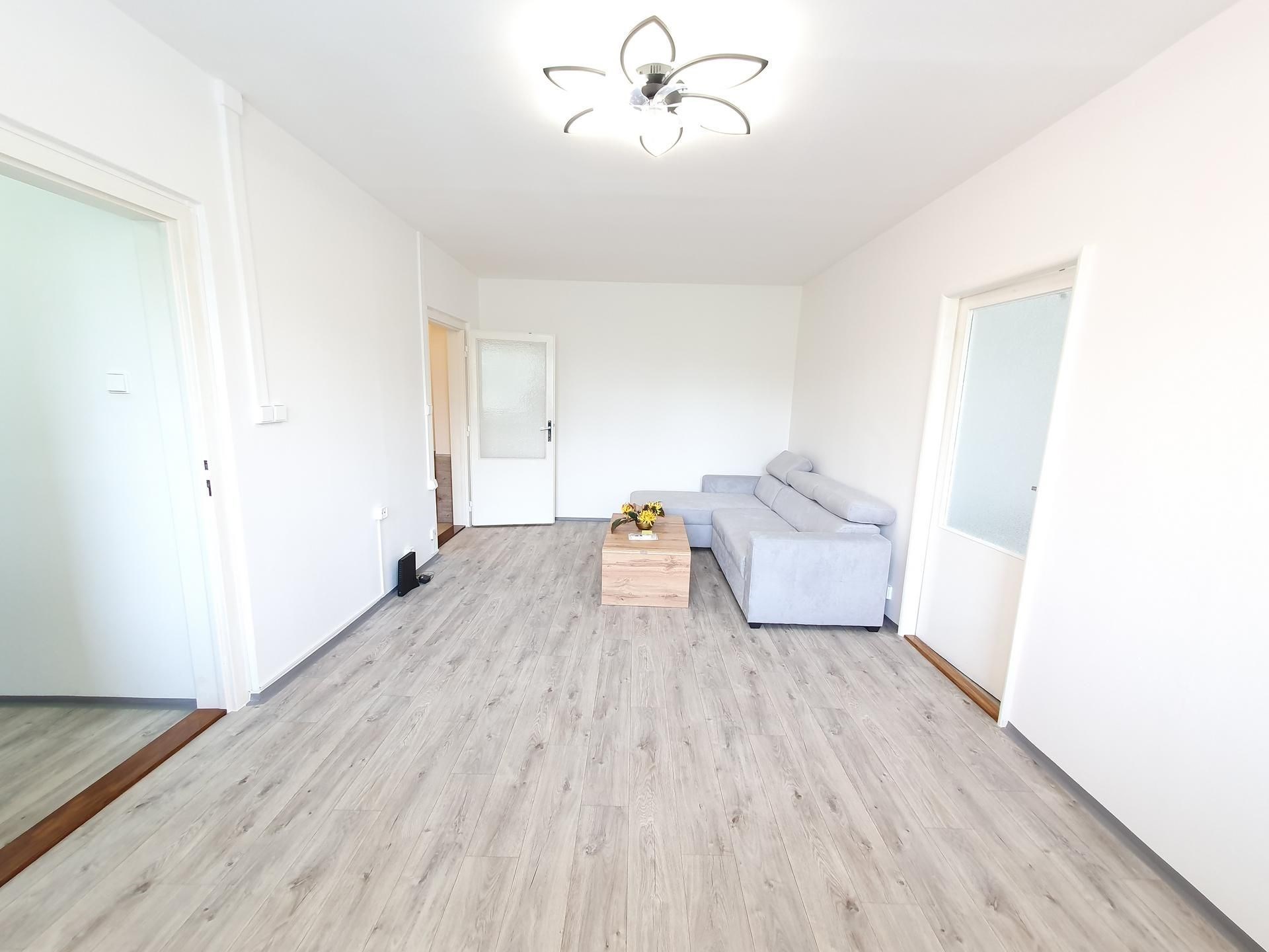 Světlý obývací pokoj s moderní parketovou podlahou, velká okna zajišťují dostatek přirozeného světla, minimalistický styl a svěží interiér
