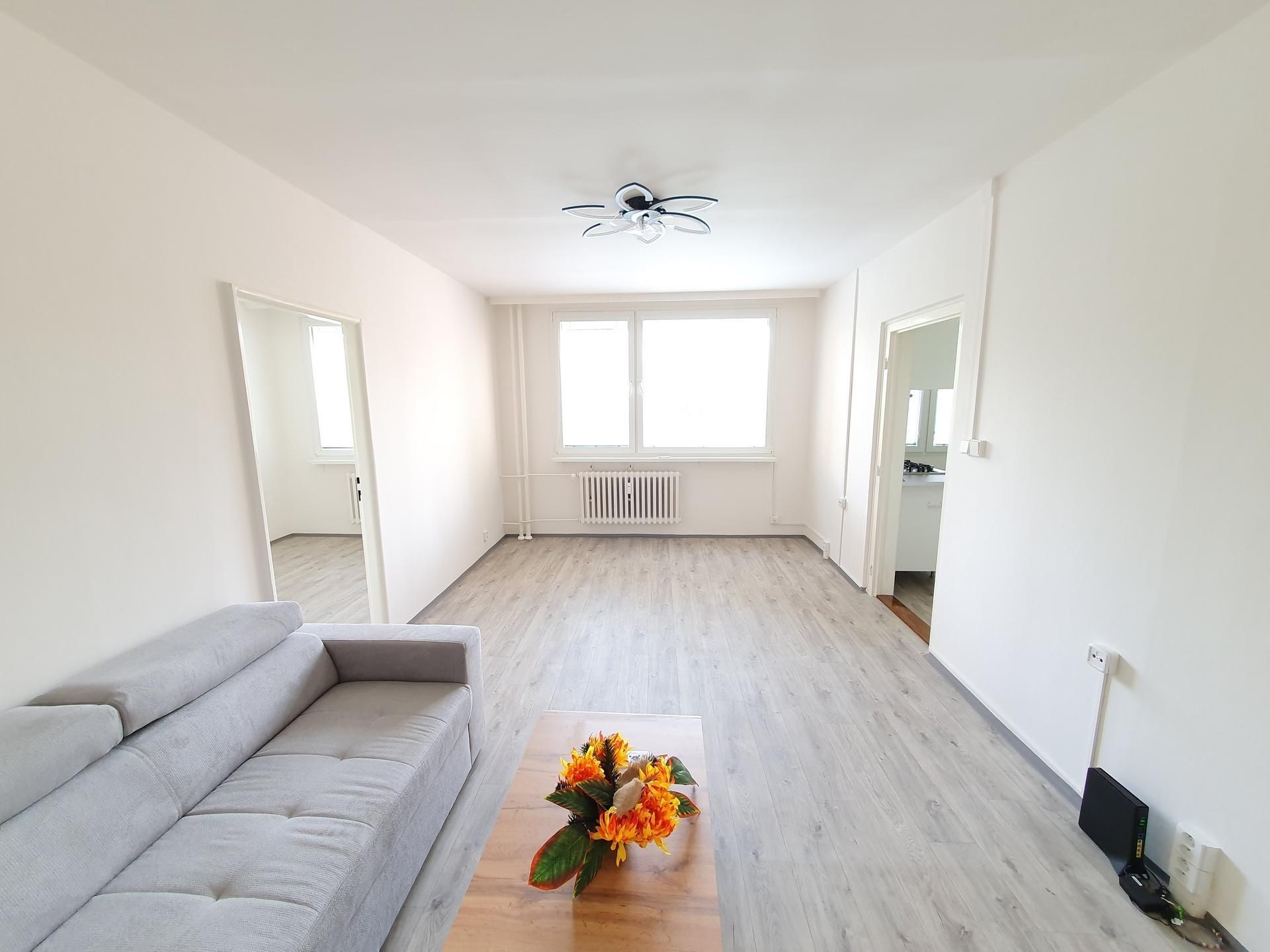 Světlý obývací pokoj s moderní plovoucí podlahou, velkými okny a minimalistickým stylem, ideální pro pohodlné bydlení.