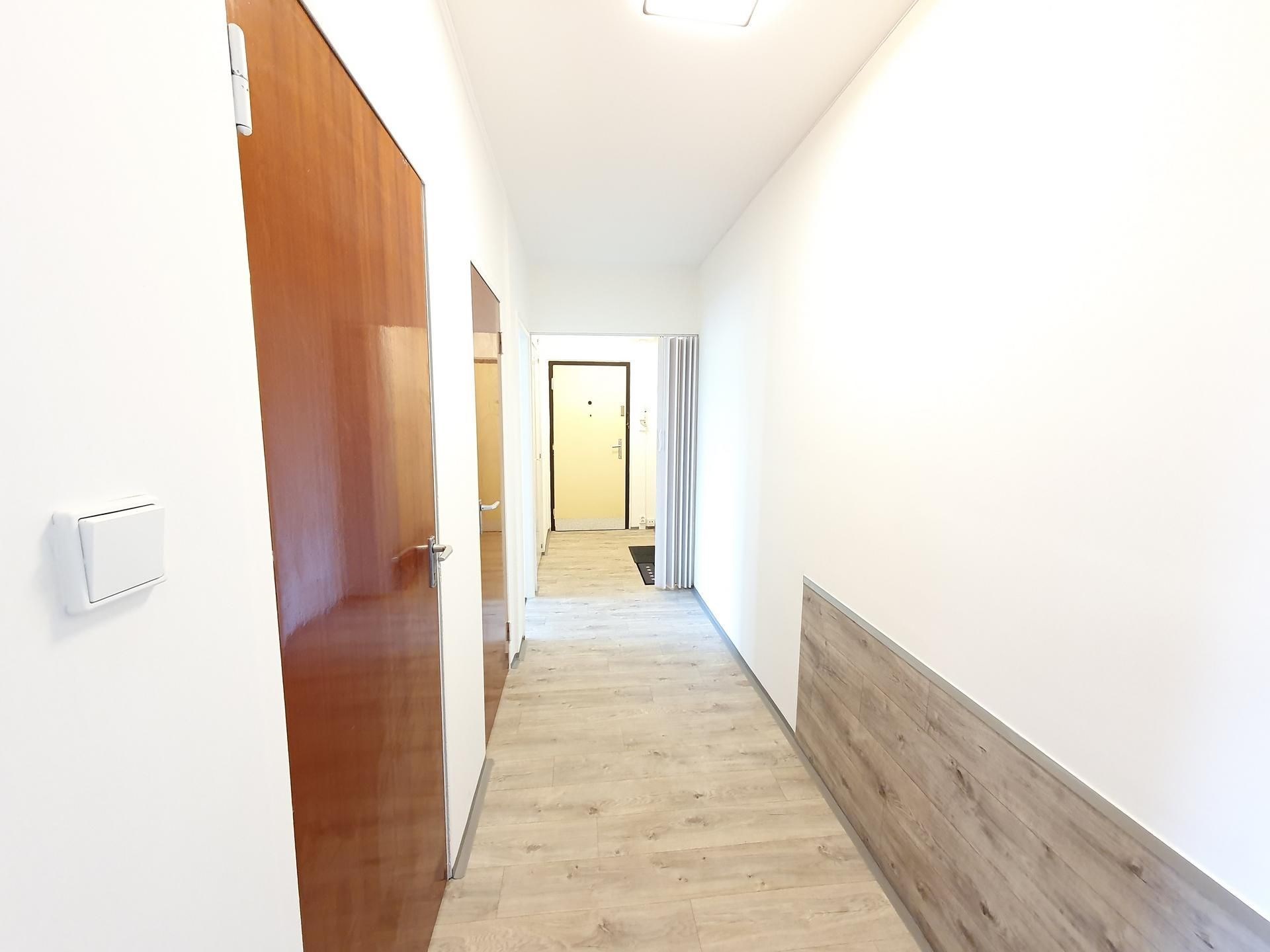 Světlá chodba s moderní laminátovou podlahou, bílými stěnami a dřevěnými dveřmi. Ideální prosvětlený prostor s minimalistickým designem.