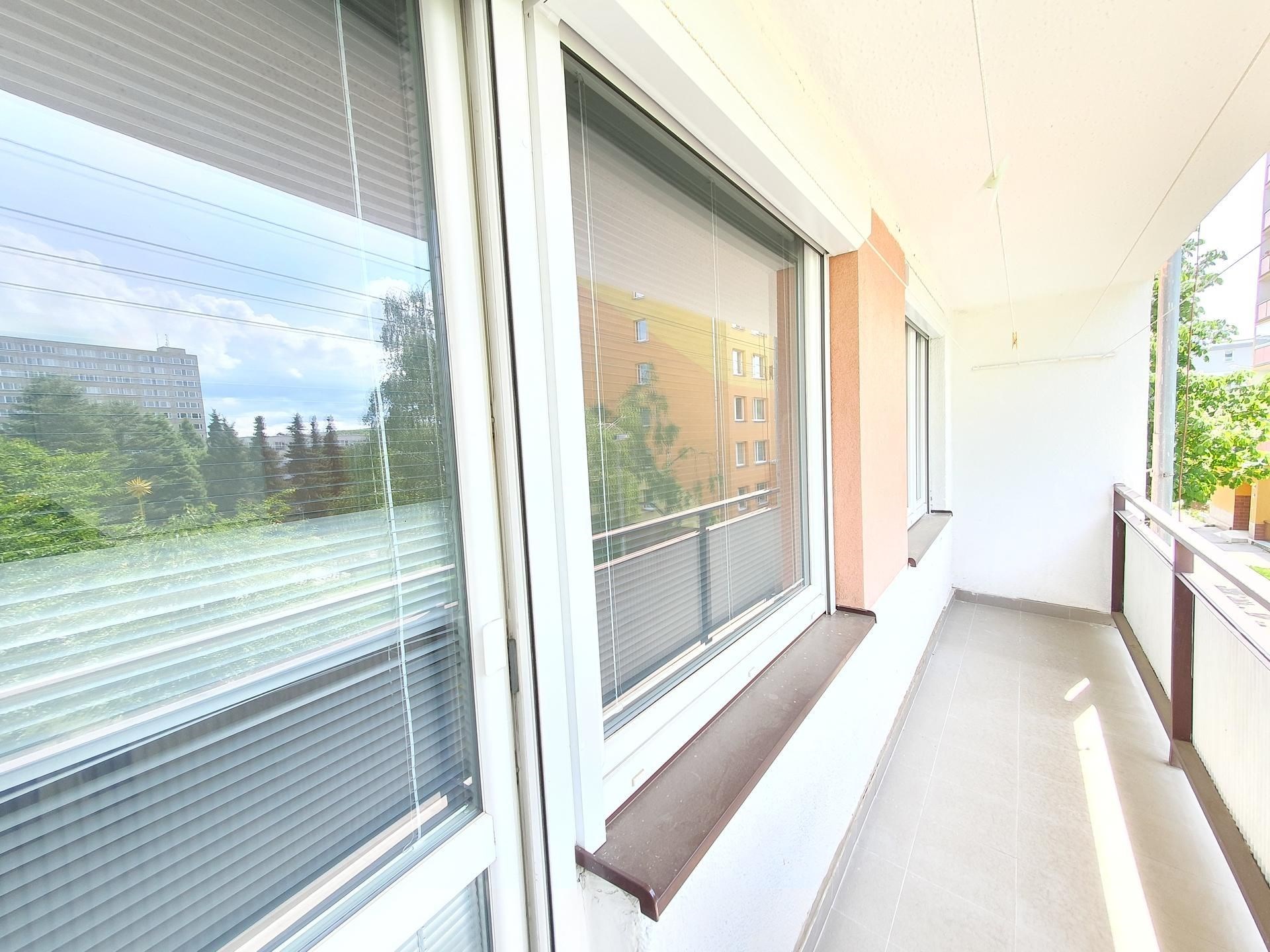 Prostorný balkon s dlažbou, velkými okny a výhledem na zelené okolí doplňuje moderní architekturu bytového domu.